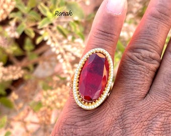 Può includere: Un anello d'oro con una grande pietra preziosa rossa di forma ovale incastonata in una montatura d'oro. L'anello è circondato da una fila di piccole pietre preziose bianche.