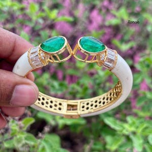 Könnte beinhalten: Ein goldfarbenes Gelenkarmband mit einem weißen Emailleband. Das Armband hat zwei große, birnenförmige grüne Edelsteine und rechteckige klare Steine. Das Armband wird von einer Hand gehalten, mit dem Namen "Ronak" im Hintergrund.