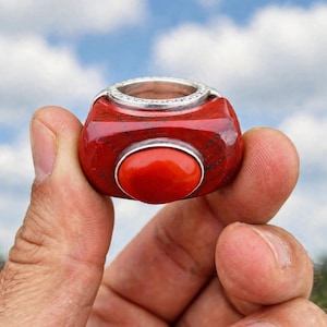 May include: A chunky ring featuring a deep red, polished stone base with dark veining and a smooth, bright orange oval cabochon set in the center. The top of the ring is finished with a silver-toned metal band featuring a textured, beaded edge.