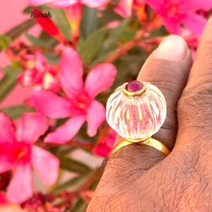 May include: A clear glass ring with a gold band and a red gemstone in the center. The ring is set against a background of pink flowers.