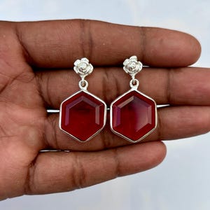 May include: A pair of silver-toned earrings. Each earring features a red, hexagonal gemstone pendant. The pendants are suspended from a floral design at the top. The earrings are displayed against a neutral background.