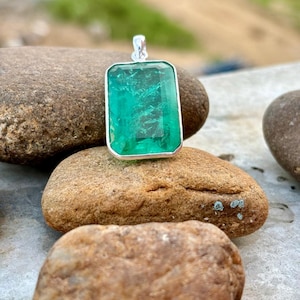 May include: A silver pendant featuring a large, rectangular, emerald green gemstone. The pendant is resting on a smooth, brown rock.