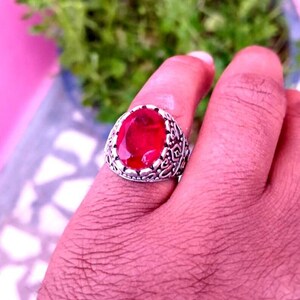 May include: A silver ring with an oval, deep red gemstone. The ring has an ornate, detailed design around the gemstone. The ring is on a finger, with a blurred green and pink background.