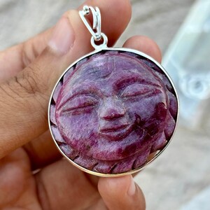 Pode incluir: Um pingente redondo com um rosto esculpido no centro, rodeado por uma moldura de prata. O rosto é esculpido em uma gema rosa e roxa.