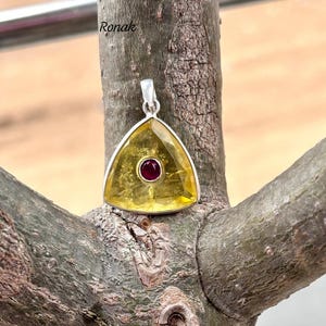 May include: A triangular pendant made of yellow citrine with a small red gemstone in the center. The pendant is set in silver and is hanging from a branch.