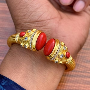 Pode incluir: Uma pulseira dourada com duas grandes pedras vermelhas ovais em cabochão nas extremidades abertas. A superfície metálica é decorada com pedras vermelhas menores e cristais transparentes brilhantes ao longo da banda texturizada.