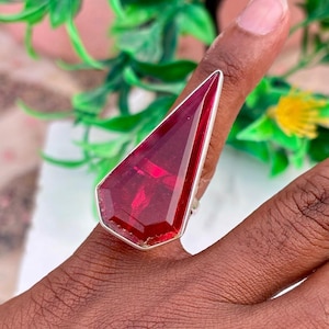May include: A large, geometric ring featuring a faceted, deep red gemstone set in a silver-toned metal. The ring's design is angular and eye-catching, with a bold, statement-making presence. The ring is shown on a finger.