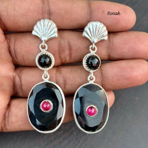 May include: A pair of silver earrings with a seashell design at the top. Each earring features a black gemstone, a smaller black gemstone, and a large oval-shaped black gemstone with a small red gemstone in the center. The earrings are designed with a drop style.
