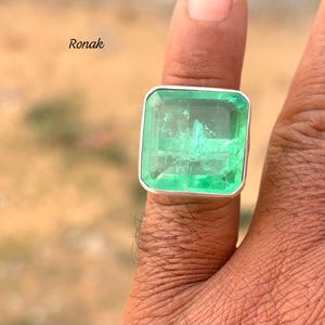 Peut inclure: Une bague en argent avec une grande pierre précieuse carrée de couleur vert émeraude. La bague est portée sur le doigt d'une personne.