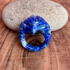 May include: A carved lapis lazuli ring in shades of deep blue with white marbling. The ring features a detailed design of a bird or mythical creature. The word "Ronak" is visible in the upper right corner.