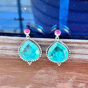 Puede incluir: Un par de pendientes plateados con una piedra preciosa de color turquesa en forma de lágrima. Cada pendiente tiene una pequeña piedra preciosa rosa en la parte superior. Los pendientes se muestran sobre una superficie de madera con el nombre "Ronak" visible.