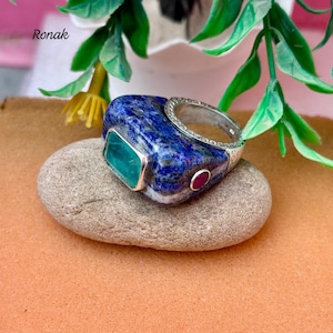 Puede incluir: Un anillo de plata con una gran piedra rectangular de lapislázuli azul, una piedra de color esmeralda y una pequeña piedra roja. El anillo está colocado sobre una piedra gris sobre un fondo color melocotón. La palabra "Ronak" es visible.