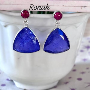 Peut inclure: Une paire de boucles d'oreilles en argent avec une pierre précieuse bleue triangulaire et une petite pierre précieuse rose au sommet. Les boucles d'oreilles sont sur une surface blanche.