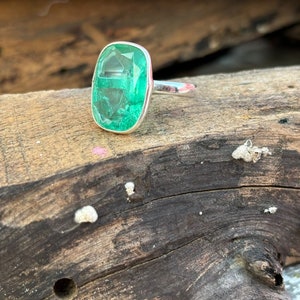 Puede incluir: Un anillo de plata con una gran gema rectangular de color verde esmeralda. La piedra está engastada en un bisel simple, resaltando su color y claridad. El anillo se exhibe sobre un trozo de madera envejecida.
