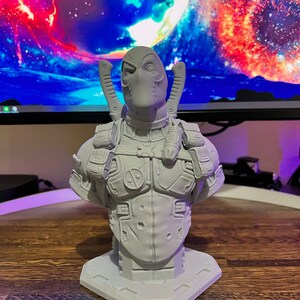 May include: A white 3D printed bust of Deadpool, a superhero character, wearing a mask and a detailed costume. The bust is on a small base.