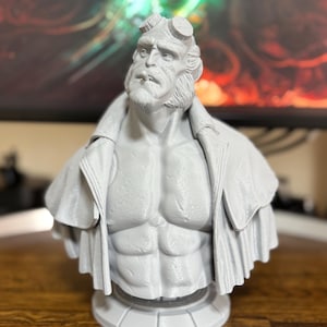 May include: A gray bust of the comic book character Hellboy, featuring detailed facial features, horns, and a coat. The sculpture is mounted on a circular base, showcasing the character's muscular physique and iconic appearance.