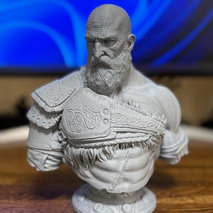 May include: A gray bust sculpture of a bearded man with a stern expression. The sculpture features detailed armor with intricate patterns and a textured fur-like chest piece. The bust is mounted on a circular base.