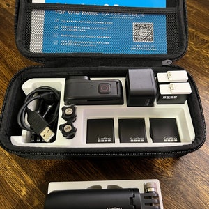 Puede incluir: Una c&aacute;mara GoPro negra con una funda negra, tres bater&iacute;as, un cable de carga y un peque&ntilde;o micr&oacute;fono negro. La funda tiene un interior blanco con compartimentos para la c&aacute;mara y los accesorios. La funda tambi&eacute;n tiene un manual de instrucciones azul y blanco.