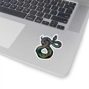 May include: A die-cut sticker featuring a coiled snake with a textured, iridescent appearance in shades of deep green, blue, and gold. The snake is depicted in a winding, S-shaped pose with a white border, adhered to the surface of a silver laptop.