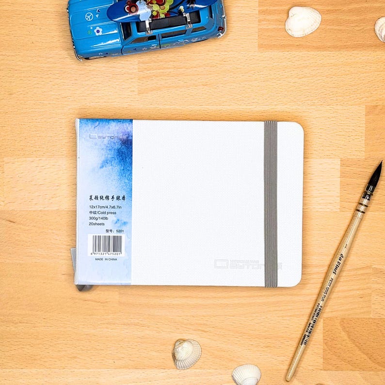 May include: A white rectangular sketchbook with a grey elastic closure band and a blue watercolour-style spine label. The label displays text including 12x17cm, 300g/140lb, and 20 sheets. A wooden paintbrush and a blue toy car are nearby.