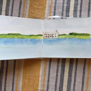 May include: An open sketchbook displaying a watercolour landscape painting. The artwork features a light blue sky, a wide expanse of blue water, a vibrant green grassy shoreline, and a small, rectangular beige building with dark windows.