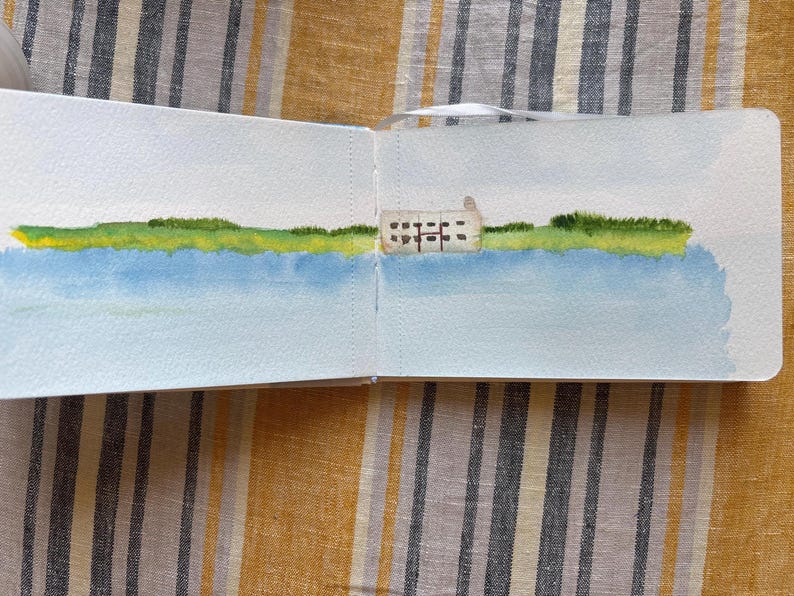 May include: An open sketchbook displaying a watercolour landscape painting. The artwork features a light blue sky, a wide expanse of blue water, a vibrant green grassy shoreline, and a small, rectangular beige building with dark windows.