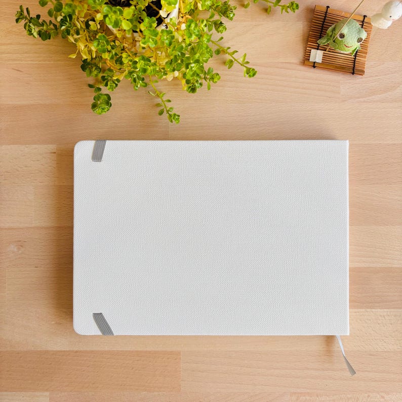 May include: A rectangular notebook with a solid white textured cover and a light grey elastic closure band positioned on the left side. The notebook features a clean, minimalist design with a white ribbon bookmark extending from the bottom edge.