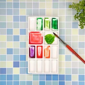 Peut inclure: Une palette de peinture aquarelle en céramique blanche avec 12 puits de peinture. La palette contient une variété de couleurs, notamment du vert, du rouge, de l'orange et du violet. Un pinceau repose sur la palette.