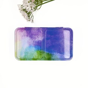 May include: A watercolour paint palette with two sections. The palette is painted with a colourful abstract design in shades of green, blue, purple, and white.