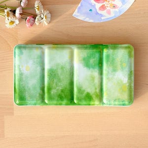 Op de afbeelding: Een groen aquarelverfpalette met drie putjes. Het palette heeft een lichtgroen en wit aquarelwasheffect.