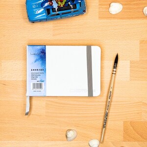 May include: A white rectangular sketchbook with a grey elastic closure band and a blue label on the left side. The label displays text including 9.5x14.5cm/3.7x5.7in, Cold press, 300g/140lb, and 20 sheets. A wooden paintbrush lies to the right.