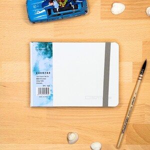 May include: A white rectangular sketchbook with a grey elastic closure band. The left side features a blue and white watercolour-style label with text including 12x17cm/4.7x6.7in, 300g/140lb, and 20 sheets. A wooden paintbrush and a blue toy car are nearby.