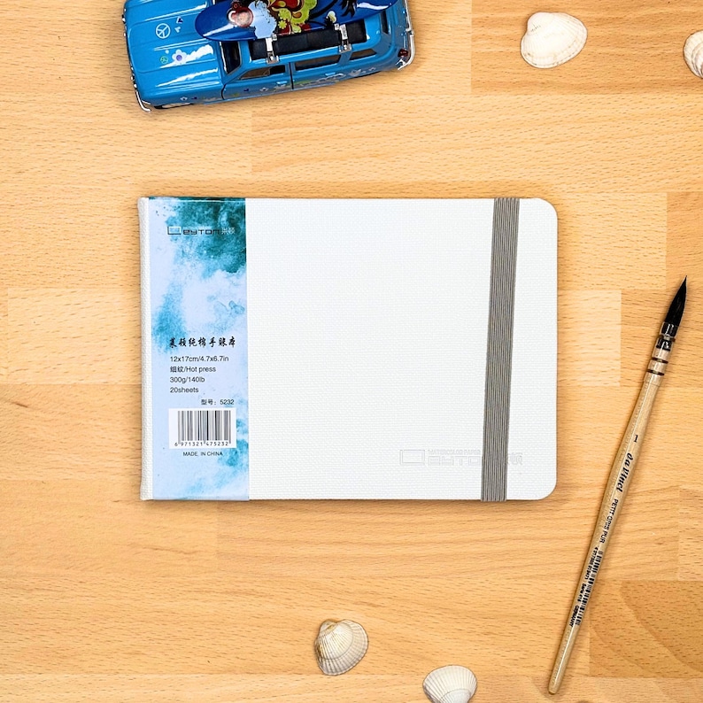 May include: A white rectangular sketchbook with a grey elastic closure band. The left side features a blue and white watercolour-style label with text including 12x17cm/4.7x6.7in, 300g/140lb, and 20 sheets. A wooden paintbrush and a blue toy car are nearby.