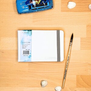 May include: A white rectangular sketchbook with a grey elastic band closure, featuring a label with the text "BAOHONG" and "300g/140lb". Beside it lies a wooden "da Vinci" paintbrush, a blue toy car with a surfboard, and several small white seashells.