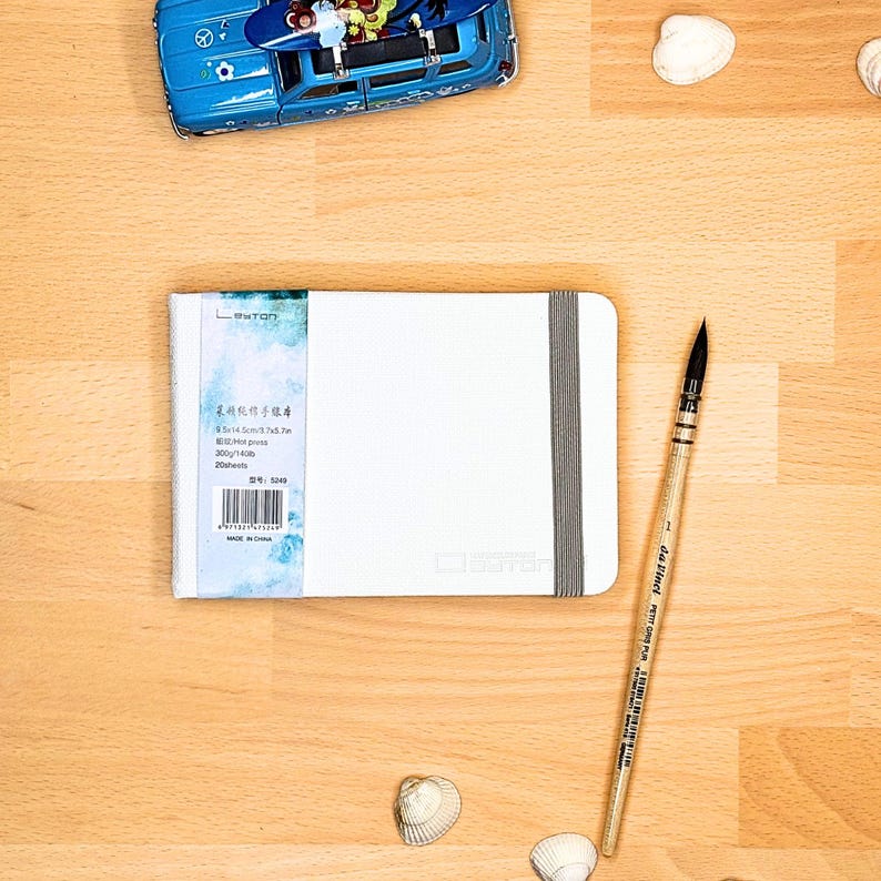 May include: A white rectangular sketchbook with a grey elastic band closure, featuring a label with the text "BAOHONG" and "300g/140lb". Beside it lies a wooden "da Vinci" paintbrush, a blue toy car with a surfboard, and several small white seashells.