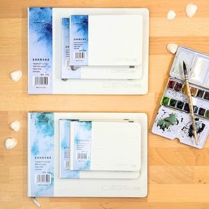 Op de afbeelding: Twee sets witte aquarelschetsboeken in verschillende formaten, elk met een blauw, aquarelachtig ruglabel met tekst en barcodes. Een metalen aquarelverfdoos met twaalf napjes verf en een houten penseel zijn inbegrepen.