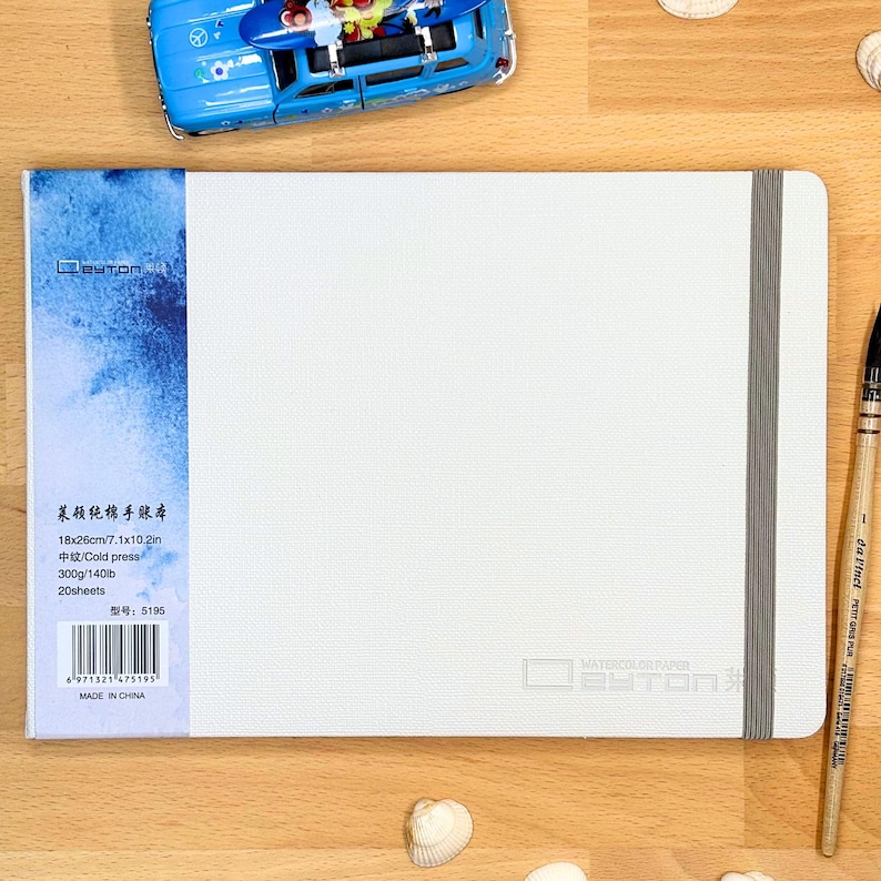 May include: A white watercolour paper sketchbook with a blue textured spine and a grey elastic closure band. The cover features the text "WATERCOLOR PAPER" and a barcode with specifications including 18x26cm, 7.1x10.2in, 300g/140lb, and 20 sheets.