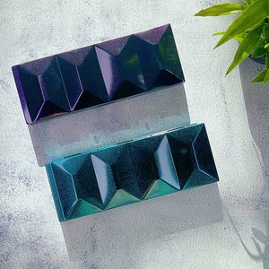 Op de afbeelding: Twee rechthoekige, iriserende make-up paletten. Het bovenste palet is dieppaars met een geometrisch ontwerp, terwijl het onderste turquoise is. Beide hebben een facetachtig, juweelachtig uiterlijk, dat licht reflecteert. De paletten liggen op een lichtgrijze ondergrond.