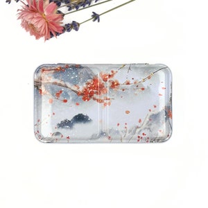 Può includere: Una scatola di latta rettangolare con un interno diviso. La scatola ha uno sfondo bianco con un disegno ad acquerello di un paesaggio innevato con fiori rossi.