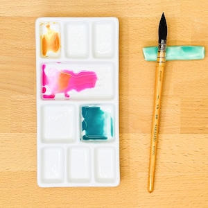 Op de afbeelding: Een witte rechthoekige aquarelpalet met verschillende vakjes met kleurrijke verf. Een penseel rust op een kleine, lichtgroene keramische houder. Het penseel heeft een houten handvat en zwarte haren. Het palet ligt op een houten oppervlak.
