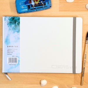 May include: A white rectangular watercolour paper sketchbook with a grey elastic closure band. The cover features a blue and white watercolour-style side panel with text including 18x26cm, 7.1x10.2in, 300g/140lb, 20sheets, and a barcode.