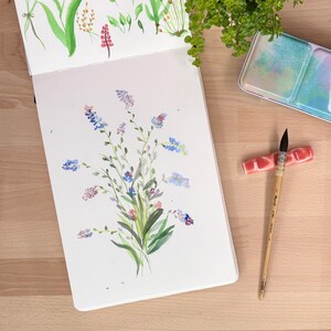 May include: An open sketchbook displaying watercolour paintings of various green leaves and small blue, purple, and pink wildflowers. Beside the sketchbook lies a wooden paintbrush with a dark tip, a pink ceramic brush rest, and a metal paint palette.