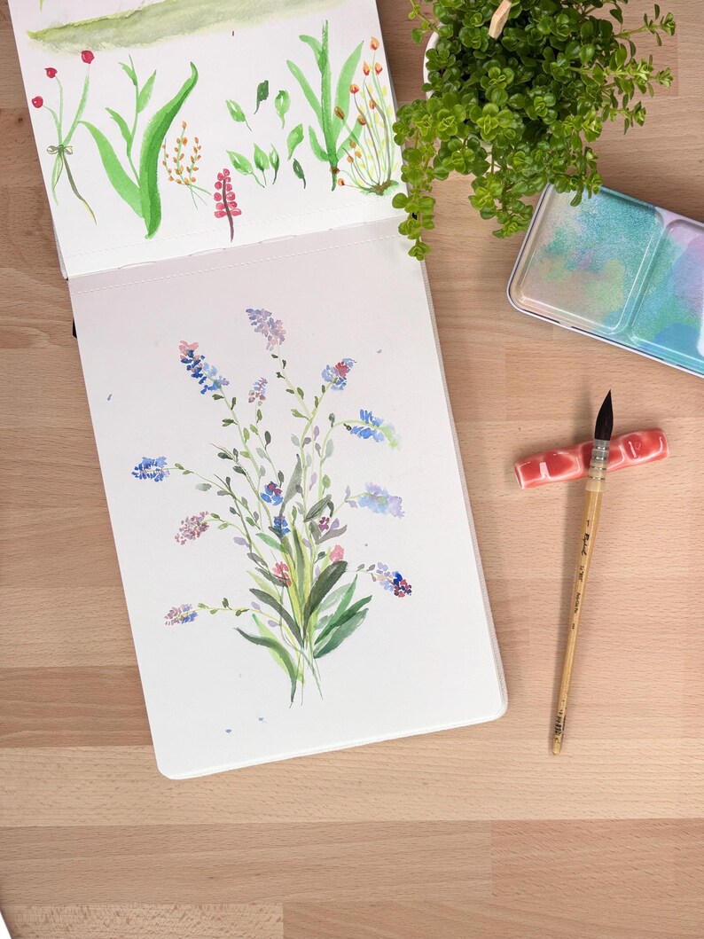 May include: An open sketchbook displaying watercolour paintings of various green leaves and small blue, purple, and pink wildflowers. Beside the sketchbook lies a wooden paintbrush with a dark tip, a pink ceramic brush rest, and a metal paint palette.