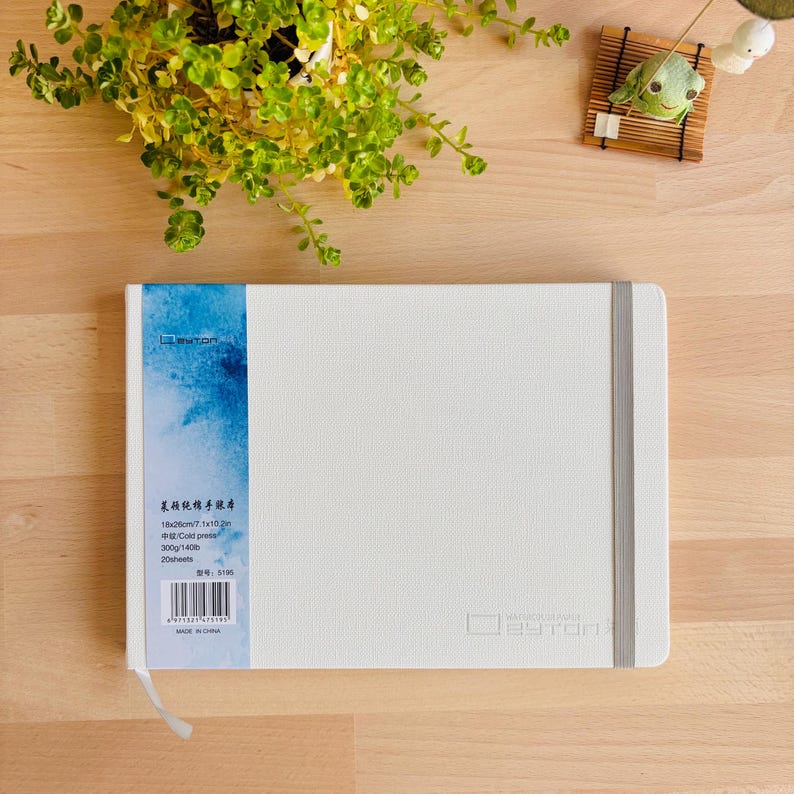 May include: A white rectangular sketchbook with a textured cover and a vertical grey elastic closure band. A blue watercolour-style paper wrap on the left side displays text including 18x26cm, 7.1x10.2in, 300g/140lb, 20 sheets, and a barcode.