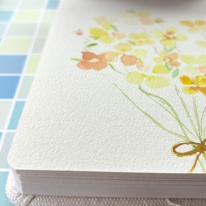 May include: A thick, white paper card featuring a watercolour illustration of a bouquet of yellow and orange flowers with green stems, tied together with a brown ribbon bow at the bottom, displayed on a textured, off-white surface.