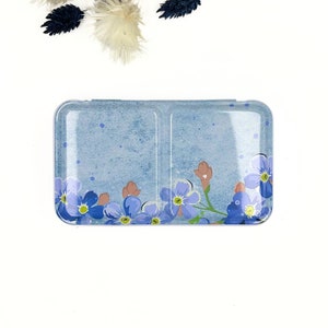Può includere: Una scatola di metallo blu con un motivo floreale. La scatola è divisa in due sezioni e presenta un motivo di fiori blu di nontiscordardime.