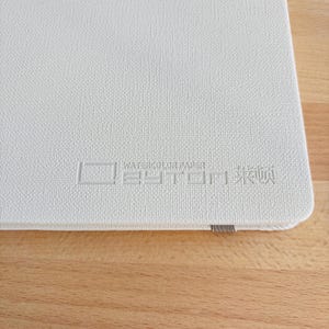 May include: A white, textured sketchbook cover featuring the embossed text WATERCOLOR PAPER and additional characters. The cover has a rounded corner and a small grey elastic band visible along the bottom edge of the rectangular notebook.