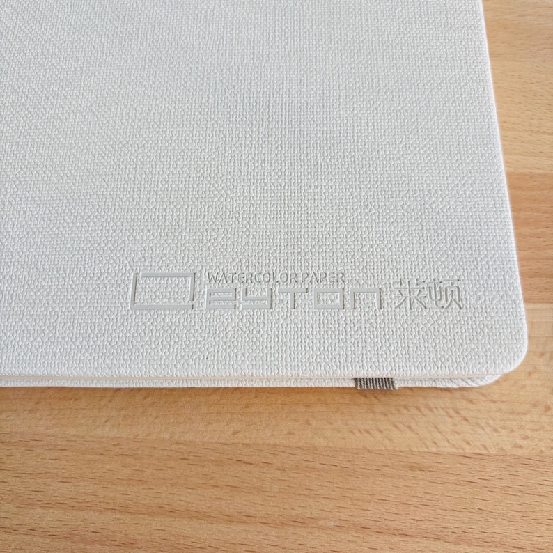 May include: A white, textured sketchbook cover featuring the embossed text WATERCOLOR PAPER and additional characters. The cover has a rounded corner and a small grey elastic band visible along the bottom edge of the rectangular notebook.