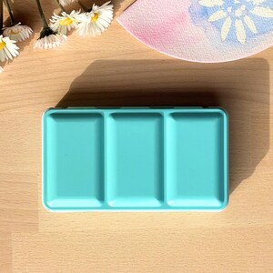 Può includere: Una tavolozza di colori ad acquerello turchese con tre pozzetti vuoti. La tavolozza è in plastica e ha una superficie liscia.