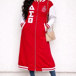 May include: A long red and white varsity jacket with a full-length zipper, white sleeves, and a white collar. The jacket features the Greek letters ΔΣΘ in white on the front and a crest on the chest. The jacket is worn with blue jeans.